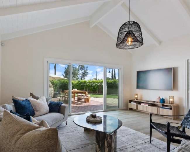 A modern living room features a large sectional sofa, a coffee table, and a wall-mounted television, with large sliding doors opening to a patio and view of greenery outside.