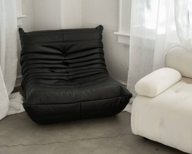 The image shows a black, contoured lounge chair next to a white, upholstered sofa in a bright, airy room with sunlight filtering through sheer curtains.