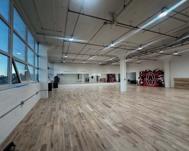 The image shows a spacious, well-lit studio with wooden flooring, large windows, and mirrors lining one wall.