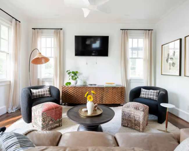 The image shows a modern living room with two armchairs, a round coffee table, a television mounted on the wall, and decorative elements including a plant and framed artwork.