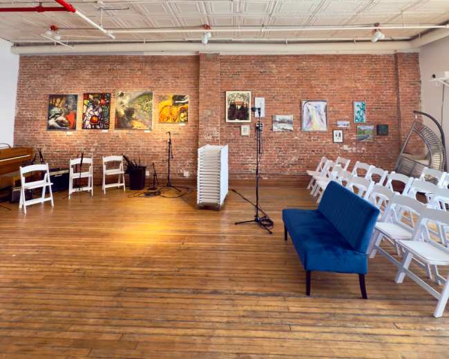 The image shows an art studio with a brick wall displaying various artworks, white chairs arranged for an audience, and a blue couch in the foreground.
