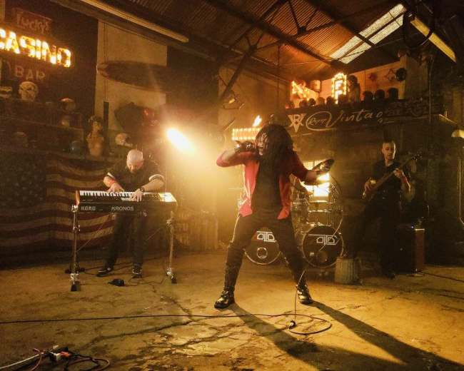 A band performs on a dimly lit stage in a rustic warehouse setting, with a keyboardist, guitarist, and a vocalist in dynamic poses.