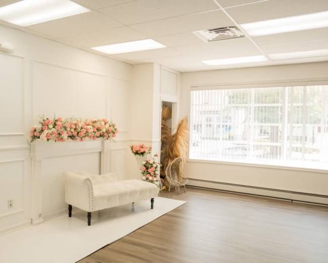 A bright, minimalist room with a white wall featuring floral decorations, a white bench, and large windows allowing natural light.
