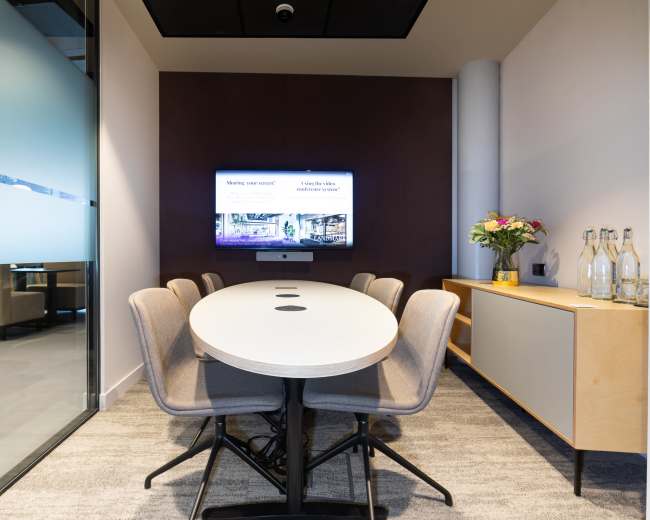 A modern conference room features a long oval table surrounded by gray chairs, with a television screen mounted on the wall displaying information, and a sideboard holding bottles of water and flowers.