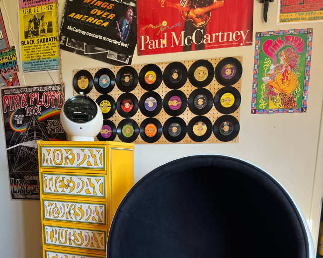 The image shows a retro-inspired room featuring a black sphere chair with a yellow blanket, a wooden chest of drawers painted in bright colors, and vinyl records displayed on the wall alongside vintage concert posters.