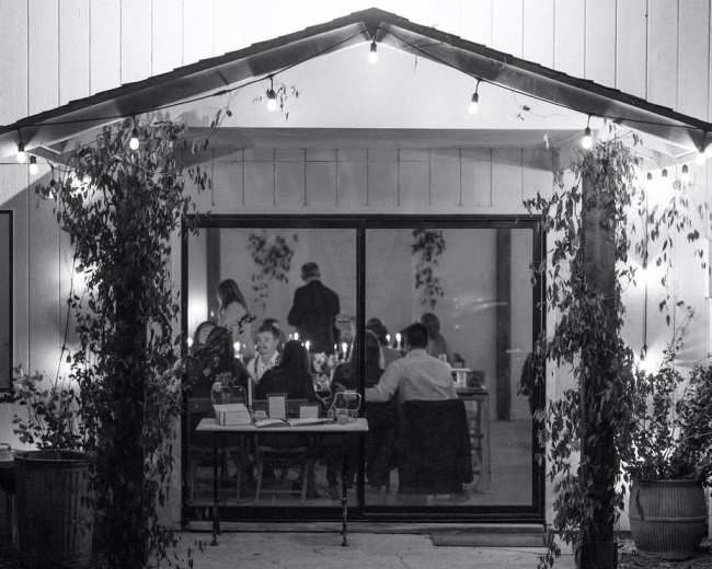 A group of people dines inside a well-lit building, visible through large glass doors framed by greenery and string lights.