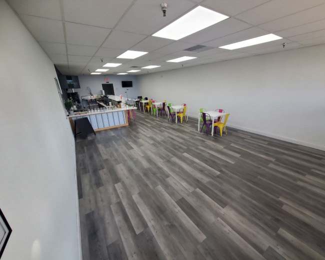 A spacious, empty room with gray wood-like flooring, white walls, colorful chairs arranged at tables along the sides, and a counter at one end.