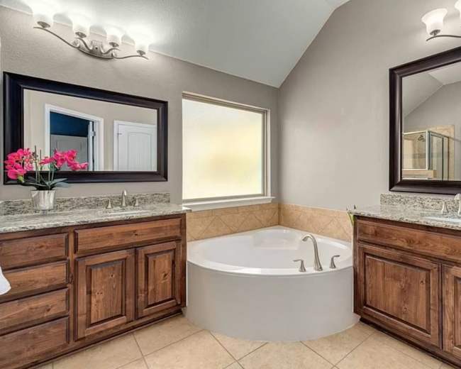 The image shows a bathroom featuring a large circular bathtub between two wooden cabinets with sinks, framed by mirrors and natural light from a window.