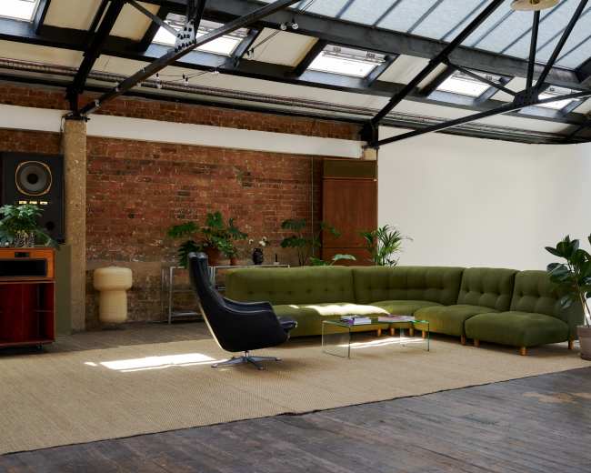 The image shows a spacious interior with a green sectional sofa, a black chair, and several plants against a backdrop of a brick wall and a white space.