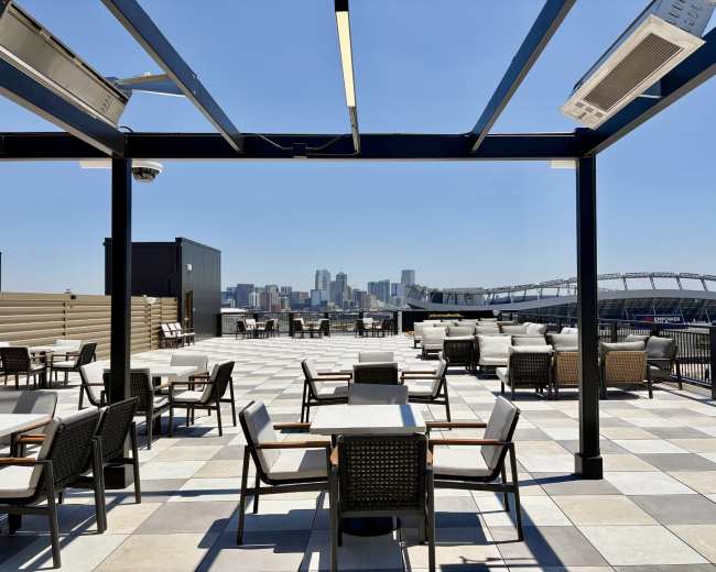 A rooftop terrace features tables and chairs with a city skyline and stadium visible in the background.