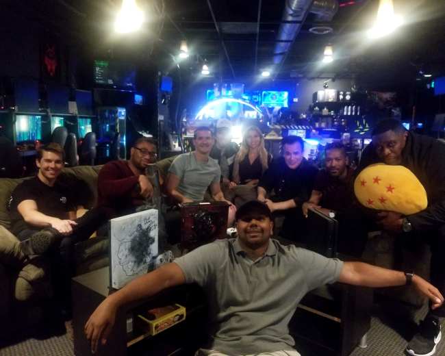 A group of eight people poses in a gaming lounge with various gaming equipment and vibrant lighting in the background.