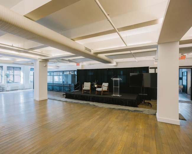 The image shows a spacious, empty event space with wooden floors, a small platform at the far end, and two chairs, along with a podium and a screen.
