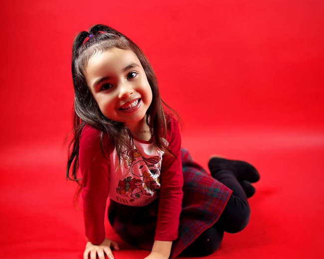 A young girl is sitting on a red background, smiling while wearing a red top and a plaid skirt.