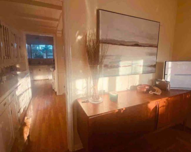 The image shows a sunlit room featuring a wooden sideboard with decorative items, leading into a kitchen with bright light streaming through the windows.