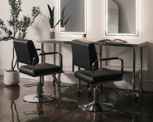 The image shows a minimalist hair salon interior featuring two mirrors above a table and two black chairs beside a potted plant.