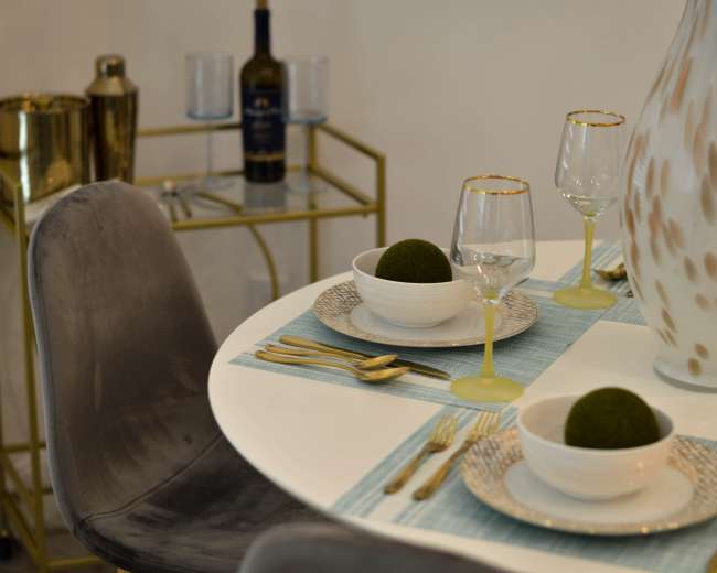 A round dining table is set with two bowls containing decorative balls, glasses, and gold-accented cutlery, with a bottle of wine and a cart in the background.