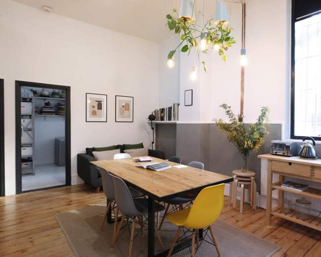 The image shows a modern dining area with a wooden table surrounded by colorful chairs, a plant hanging from the ceiling, and a large window providing natural light.