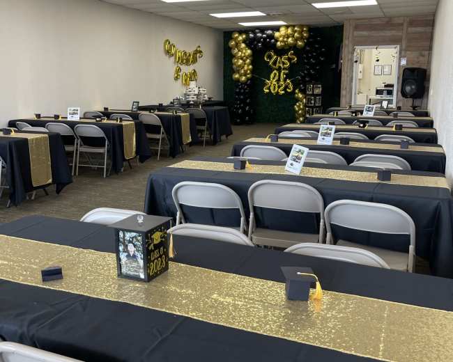 A decorated hall set up for a graduation celebration, featuring several tables with black and gold tablecloths, chairs, and balloons on the walls.