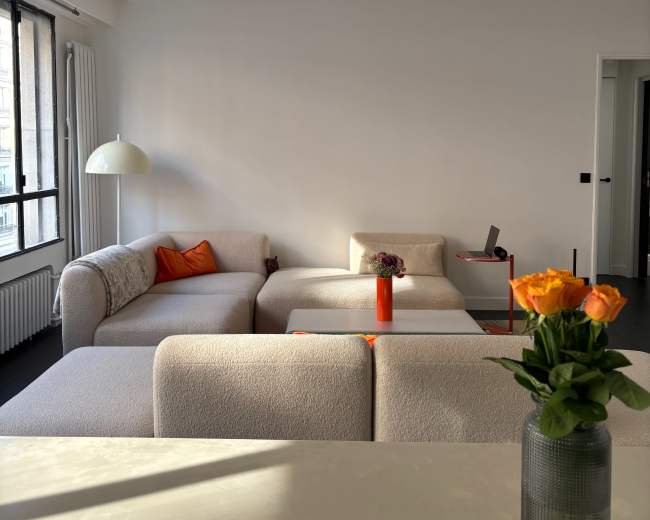 A bright living room features several light-colored, modern couches, a small table with a laptop, and a vase of orange flowers on a surface in the foreground.