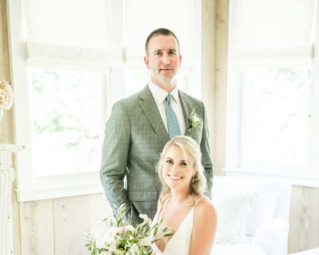 A bride in a white dress holds a bouquet while sitting on a bed, and a groom in a suit stands behind her in a well-lit room.