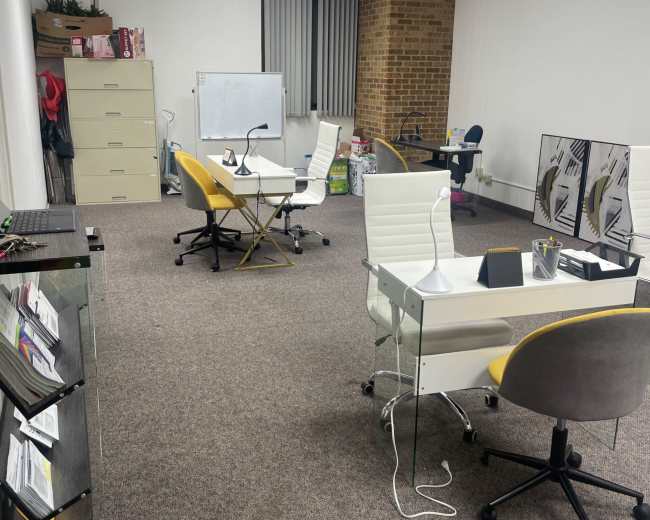 A sparsely furnished office space with two desks, a filing cabinet, and a whiteboard against the wall.