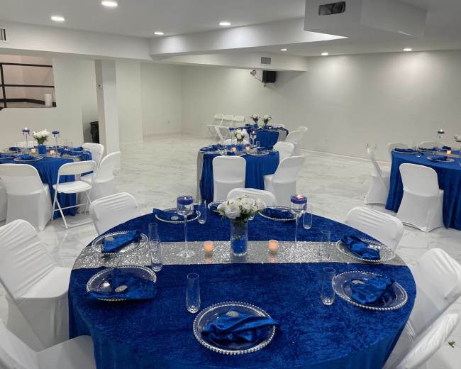 The image shows a spacious event room with white walls and marble flooring, featuring round tables covered in blue tablecloths, each set with white chairs and decorative centerpieces.