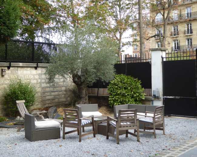 A small outdoor seating area is set with wooden chairs and a table, surrounded by gravel, plants, and an olive tree.