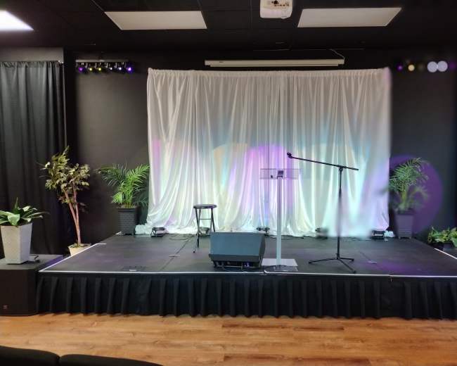 A stage with a microphone, a lectern, and potted plants, set against a backdrop of white drapery.