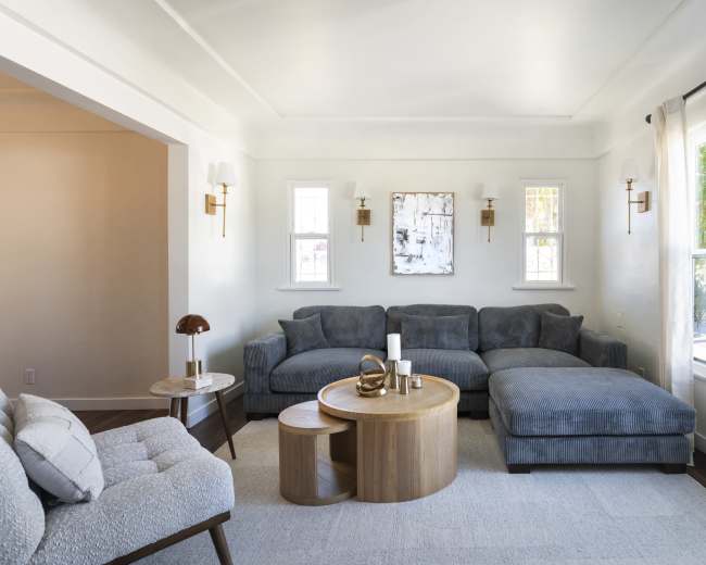 The image shows a modern living room featuring a gray sectional sofa, a round wooden coffee table, and large windows that allow natural light to fill the space.