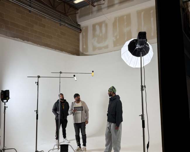 Three individuals are positioned in a photography studio surrounded by lighting equipment and a smooth white backdrop.