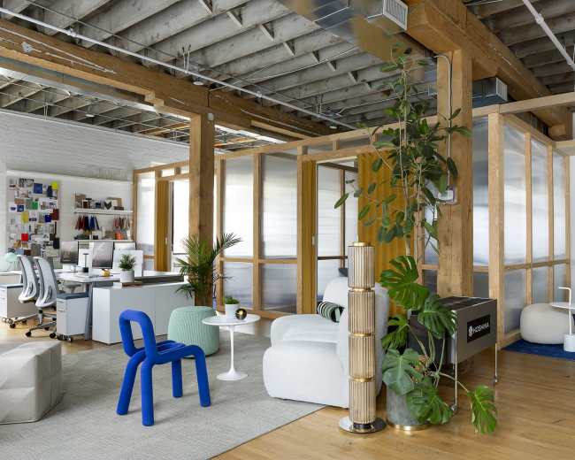 The image shows a modern office space featuring wooden beams, glass partitions, and various seating areas with plants and colorful furnishings.