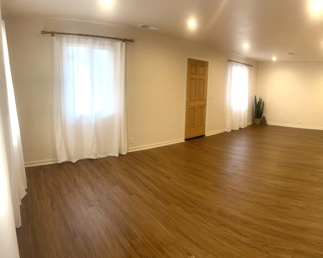 A spacious room with light-colored walls, wooden flooring, a large window with sheer curtains, and minimal furnishings.