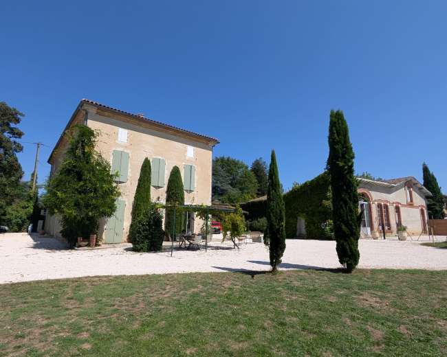 The image shows a two-story light-colored house with green shutters surrounded by neatly trimmed cypress trees and a gravel area under a clear blue sky.