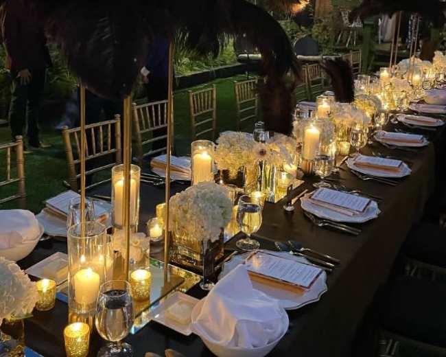 A long banquet table is set outdoors with elegant decor, surrounded by greenery and string lights.