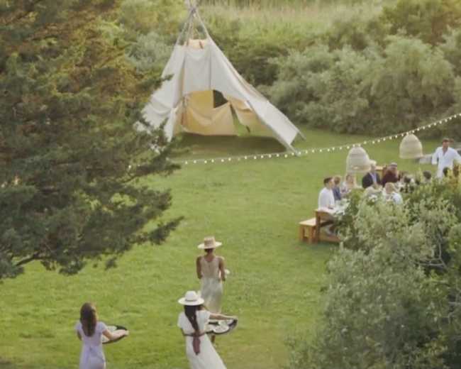 A group of people gathers in a grassy outdoor area featuring a tent, picnic tables, and decorative lights.