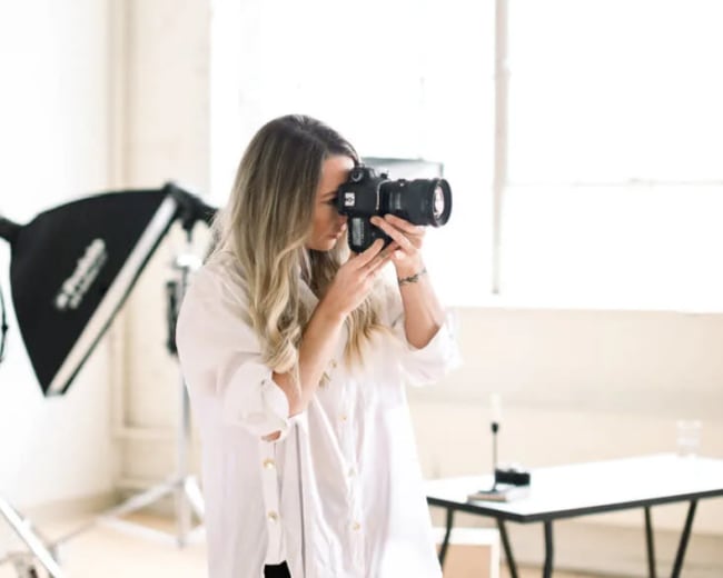 A woman is holding a camera and taking a photo in a bright, minimalist studio with large windows.