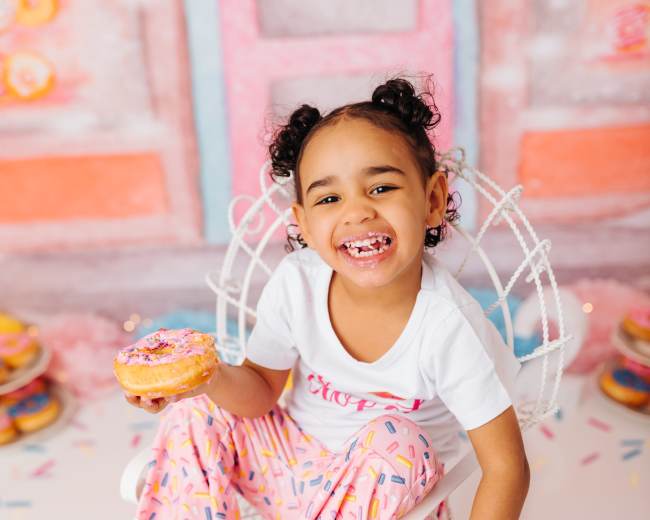 A young girl with curly hair sits in a white chair holding a donut while smiling and wearing colorful pajamas.