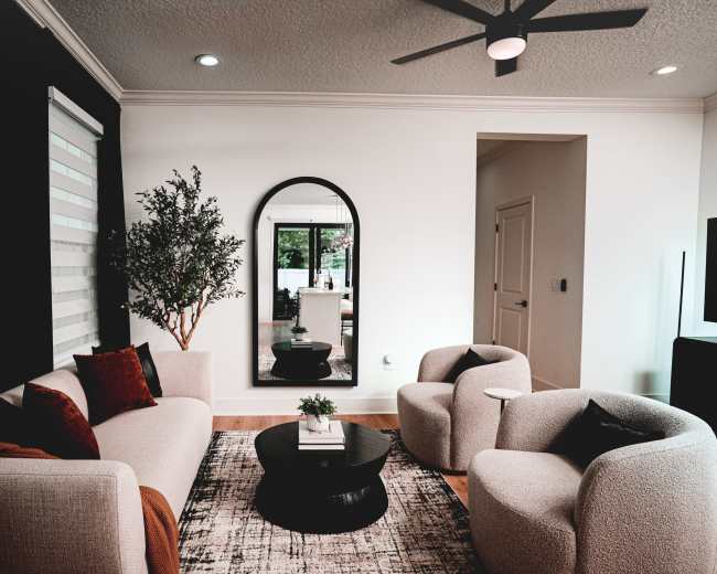 A modern living room features a sofa, two armchairs, a round coffee table, and a large mirror reflecting a light and airy space.