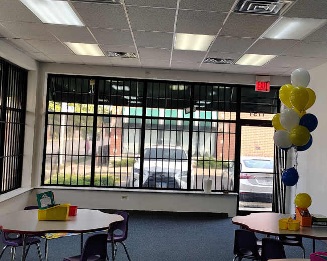 The image shows a brightly lit classroom with tables and chairs arranged, a window displaying a parked car outside, and a cluster of balloons in blue and yellow at the corner.