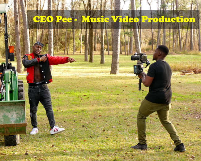 A man dressed in a red and black jacket performs while another man films him with a camera, near a woman seated on a green tractor in a wooded area.