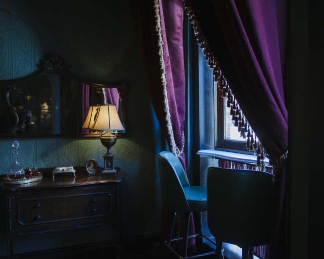 A dimly lit room features a decorative mirror, a wooden dresser, a lamp, and a window with rich curtains.