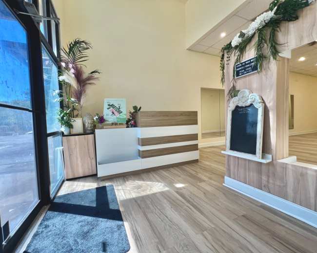 The image shows a bright, modern reception area featuring a sleek front desk, decorative plants, and a chalkboard element on the wall.