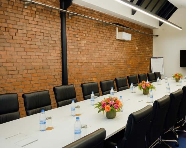 A large conference room features a long table with black chairs, decorated with floral centerpieces and water bottles, against a backdrop of exposed brick walls.