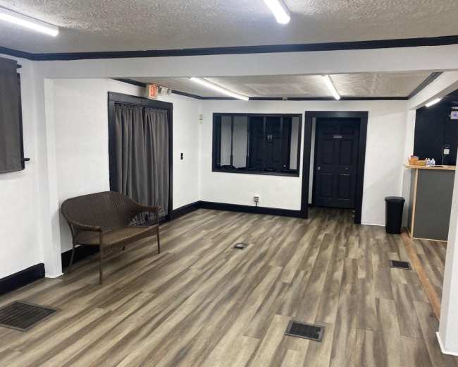 The image shows a sparsely furnished interior space with wood-like flooring, white walls, and black trim, featuring a chair, window, and an open doorway.