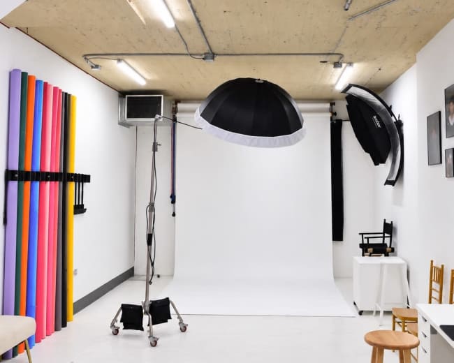 The image shows a photography studio with a white backdrop, lighting equipment suspended from the ceiling, and colorful backdrop rolls on one wall.