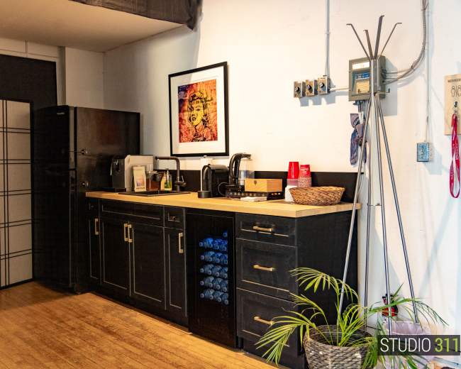 The image shows a compact kitchen area with dark cabinets, a countertop featuring a coffee maker, and a refrigerator, along with a wall-mounted shelf and a plant in the corner.