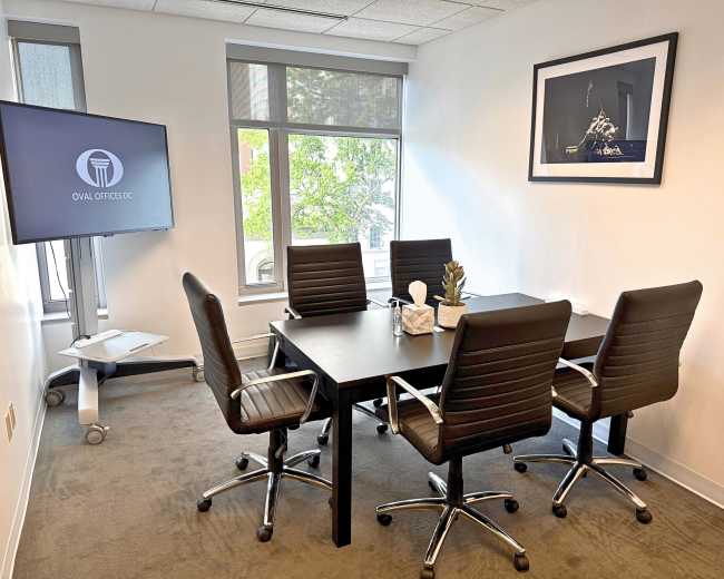 The image shows a small conference room with a table surrounded by five black office chairs, a monitor on a stand, and large windows allowing natural light.