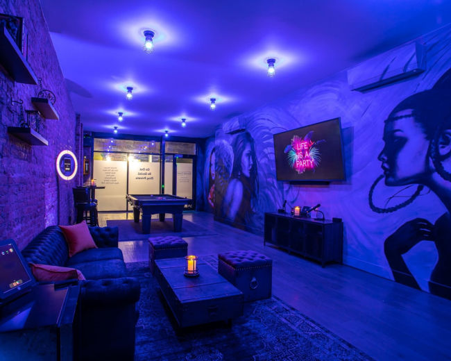 A dimly lit lounge area features brick walls adorned with murals, a pool table, seating arrangements, and a television displaying neon-themed art.