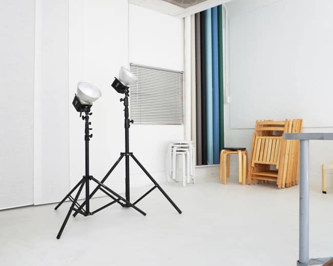 Two studio lights on tripods positioned in a minimalist room with a few chairs stacked against a wall.