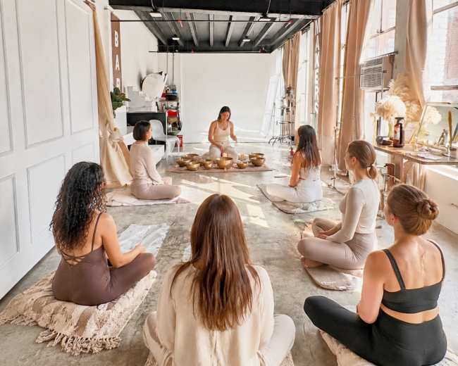 A group of seven people sit on mats in a spacious, softly lit room while a facilitator leads a session at the front.